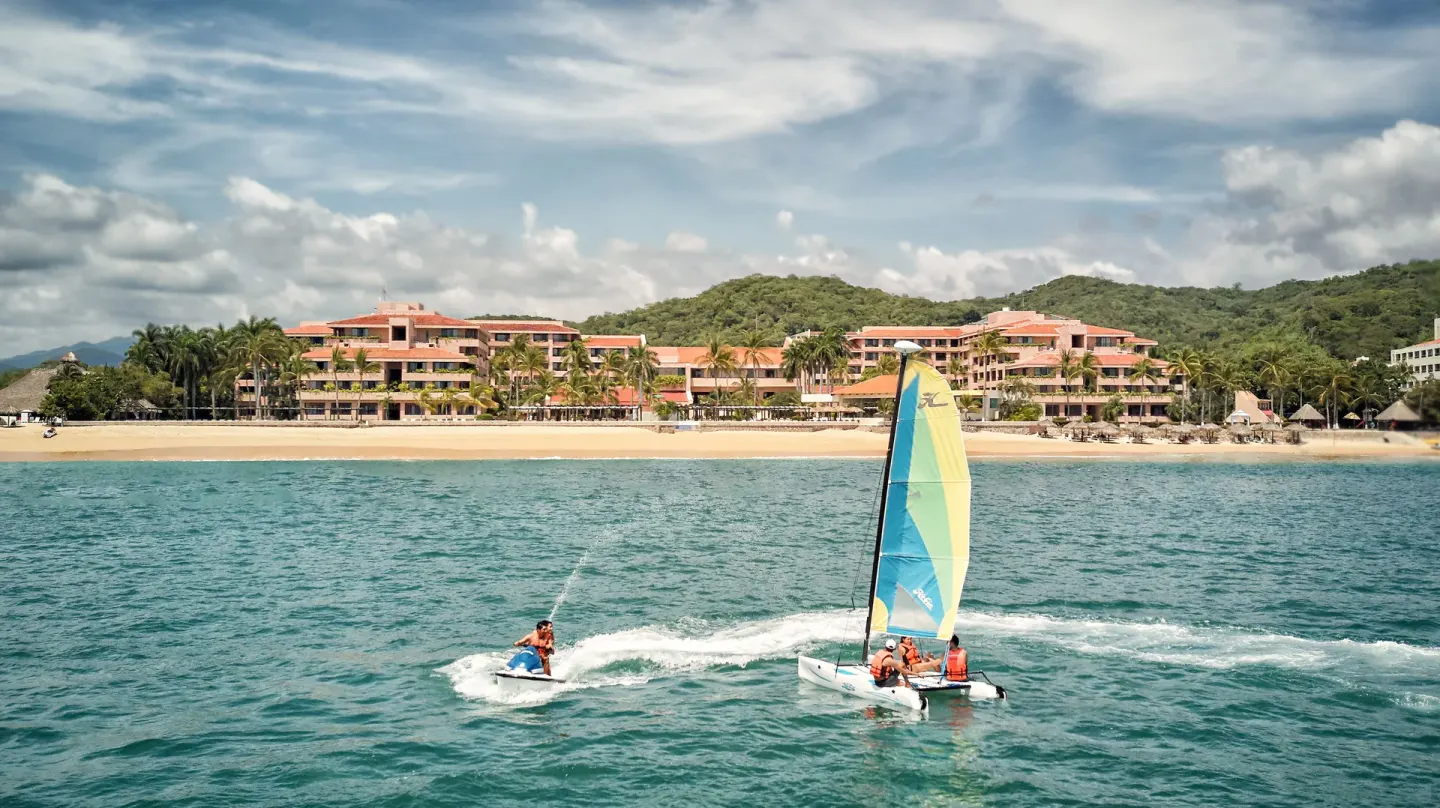 A sailboat and jet ski cross the water in front of Barceló Huatulco and the beach.