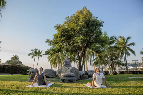 Two guests practice yoga on a lawn with palms and a statue at Barceló Karmina.