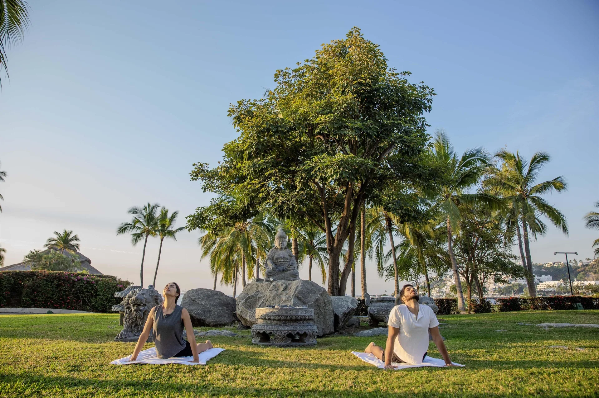 Two guests practice yoga on a lawn with palms and a statue at Barceló Karmina.