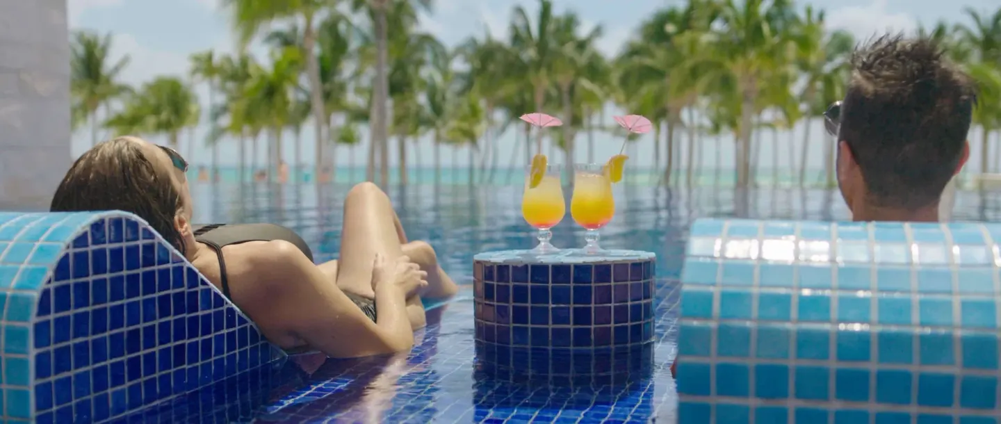 Two guests lounge in a tiled pool with tropical drinks at Barceló Maya Riviera.