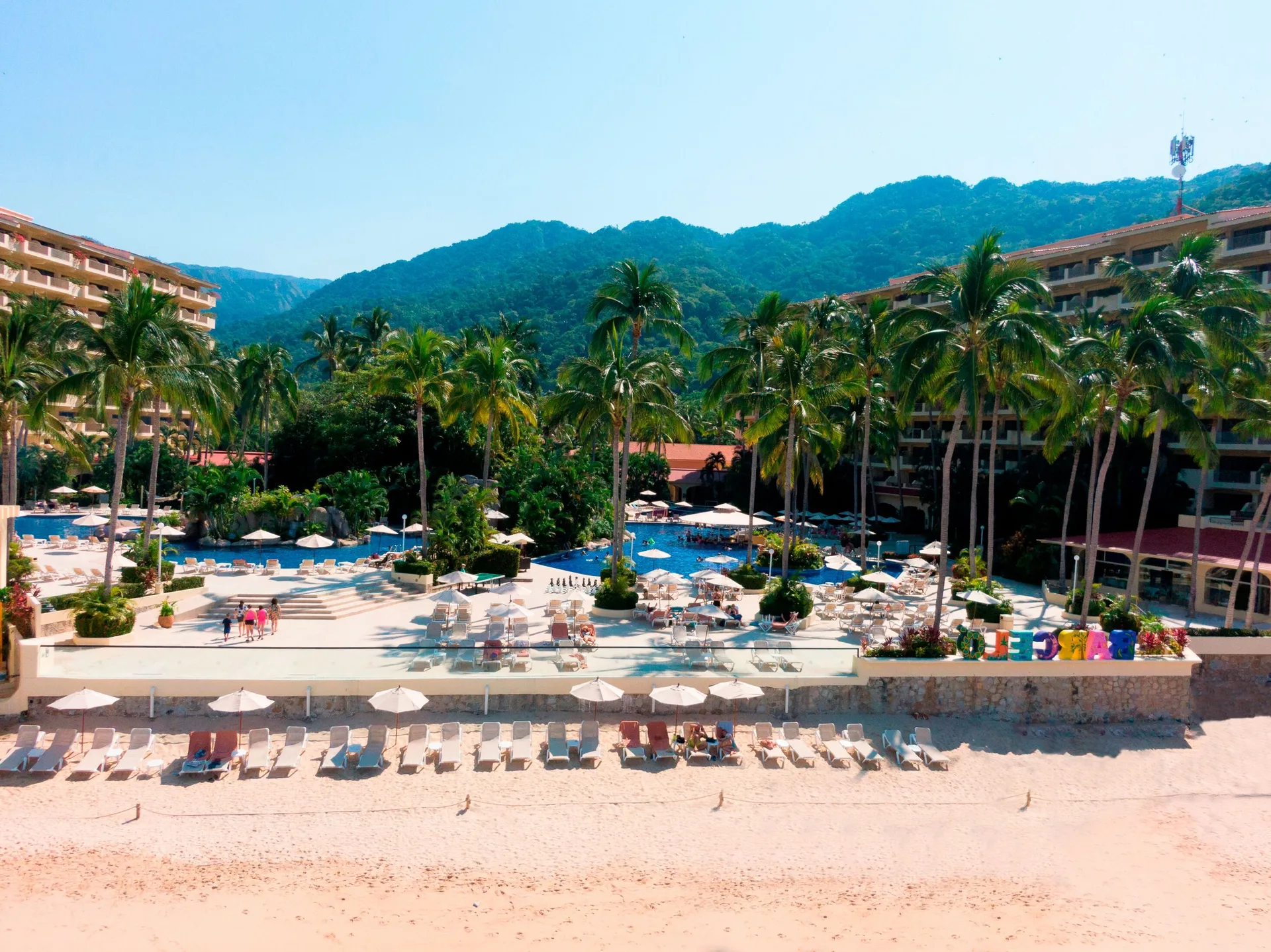Beachfront pool terrace with palms, loungers, resort buildings, and mountains at Barceló Puerto Vallarta.