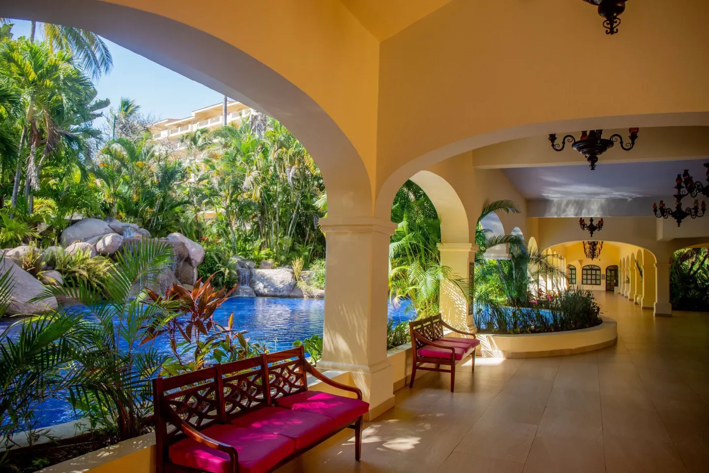 Covered garden walkway at Barceló Puerto Vallarta with benches, arches, palms, and water.