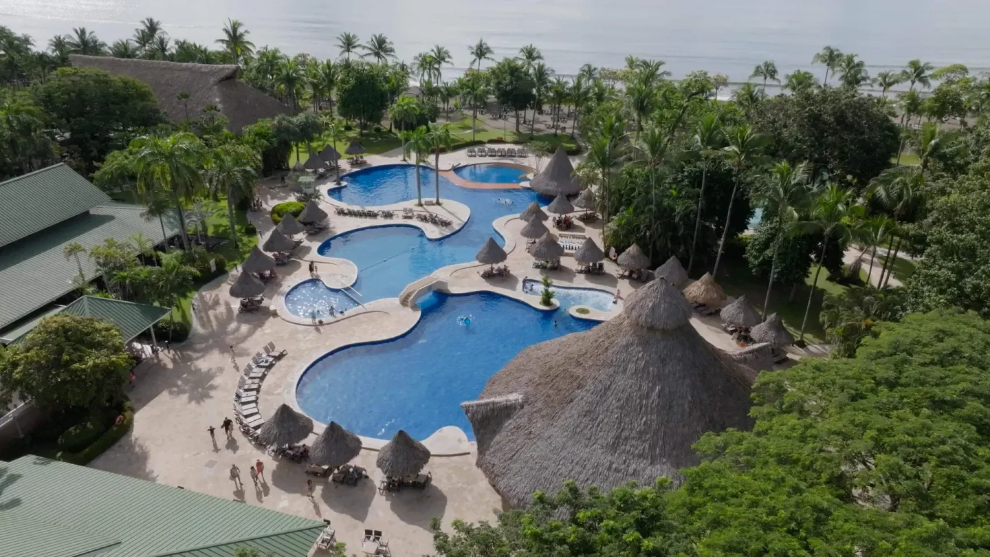Aerial view of pools, thatched umbrellas, palms, and beach beyond at Barceló Tambor.