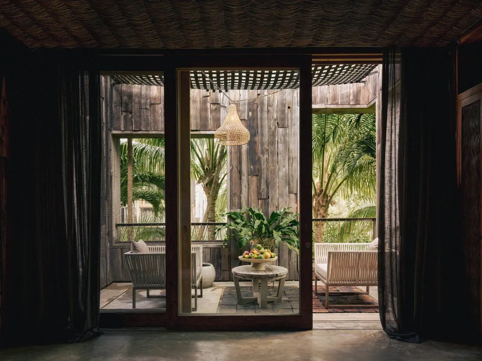 Wood-framed glass doors opening to a shaded balcony with seating and palms at BE Destination Tulum