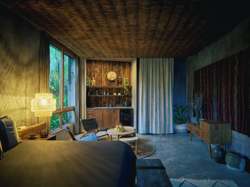 Evening view of a guest room living area with woven ceiling, seating and curated shelving at BE Destination Tulum