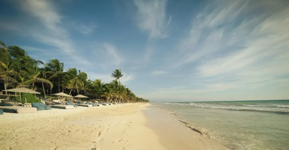 Empty stretch of beach with low loungers and palms meeting the calm sea at BE Destination Tulum