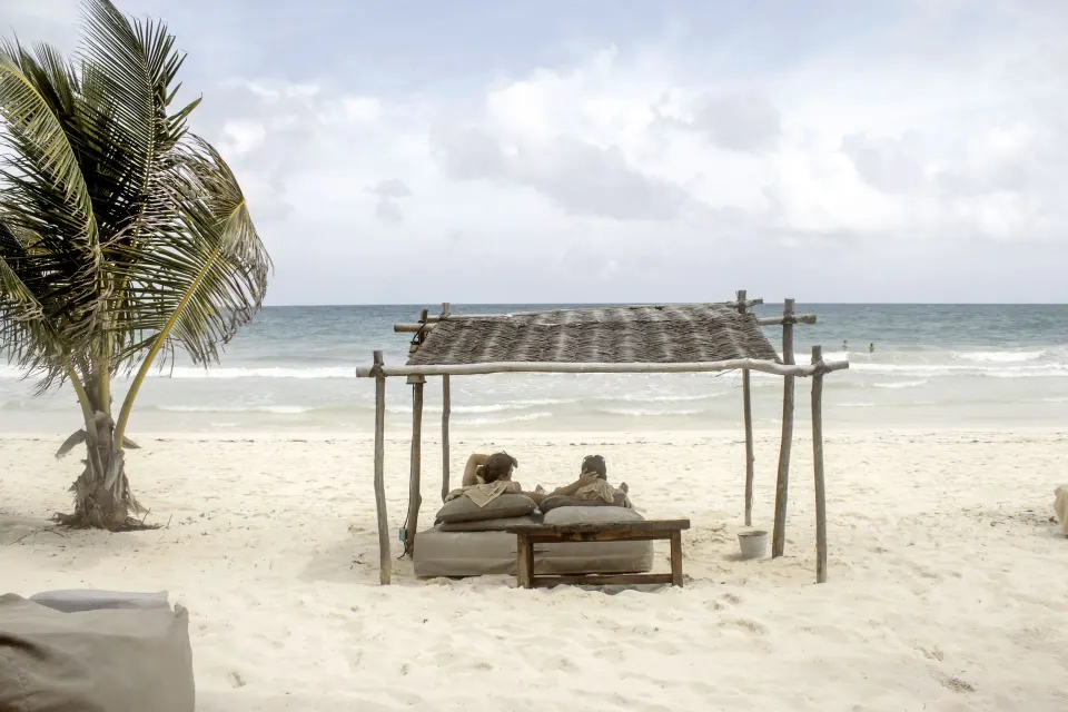 Two lounge beds under a rustic palapa on the beach at BE Destination Tulum