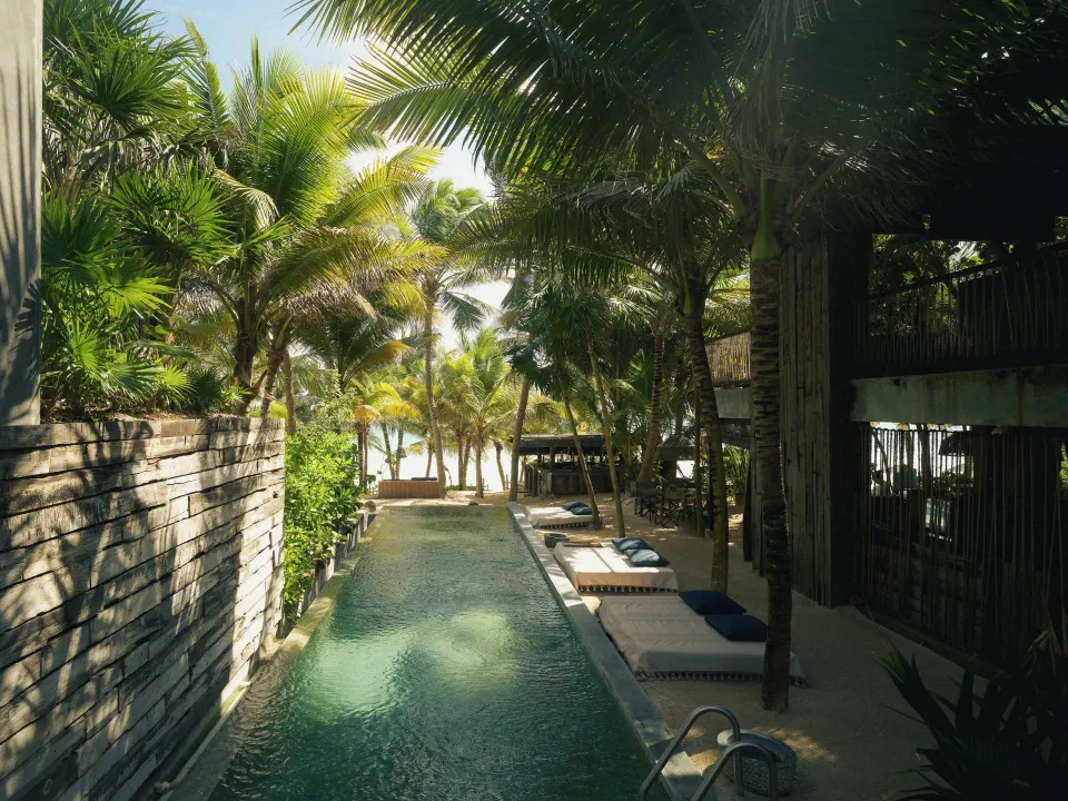 Long pool corridor lined with palms and loungers toward the beach at BE Destination Tulum
