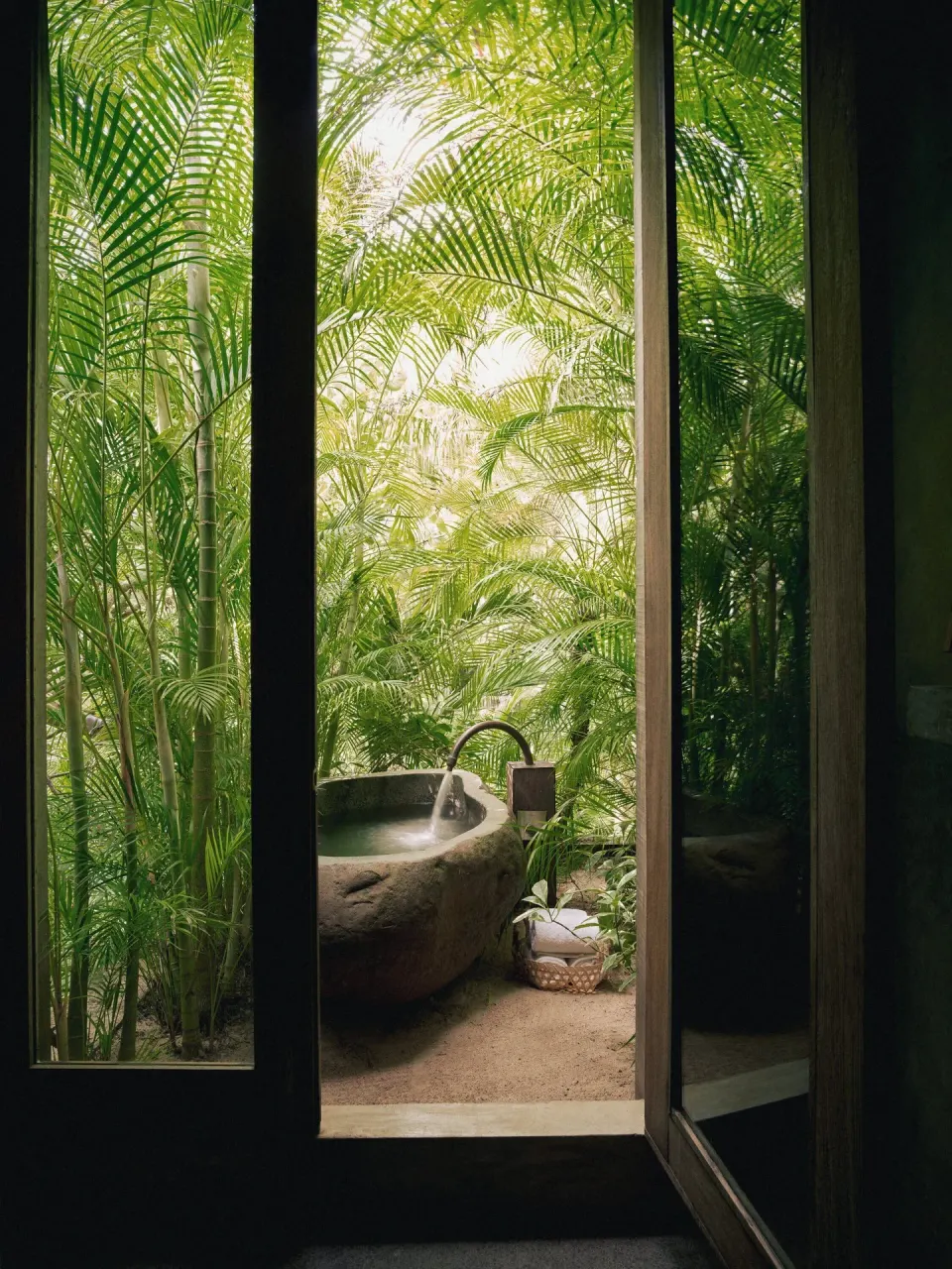 Stone outdoor bathtub surrounded by palms visible through glass doors at BE Destination Tulum