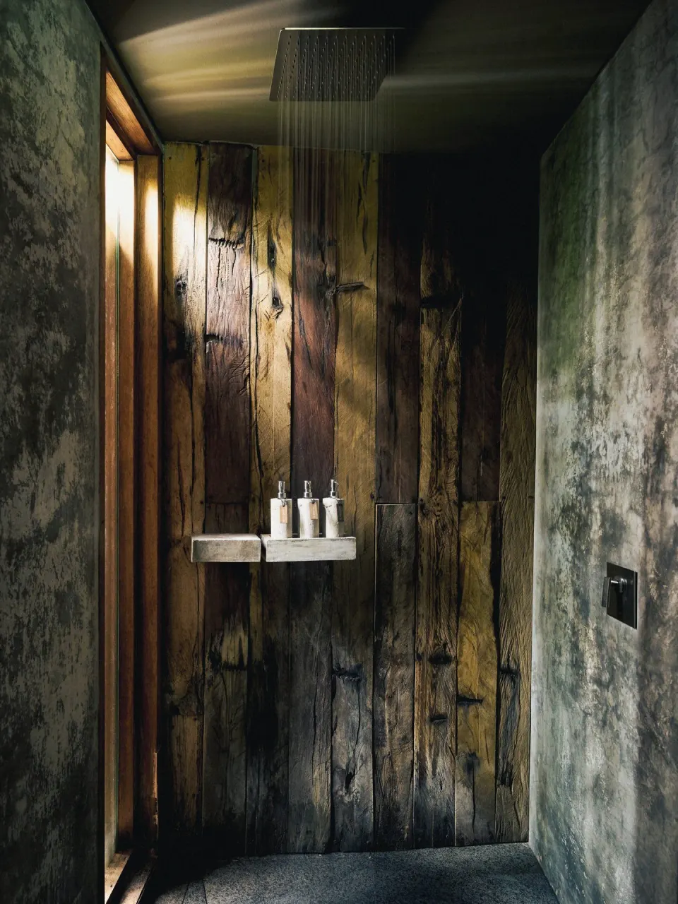 Rain shower with dark wood back wall and toiletries on a shelf at BE Destination Tulum