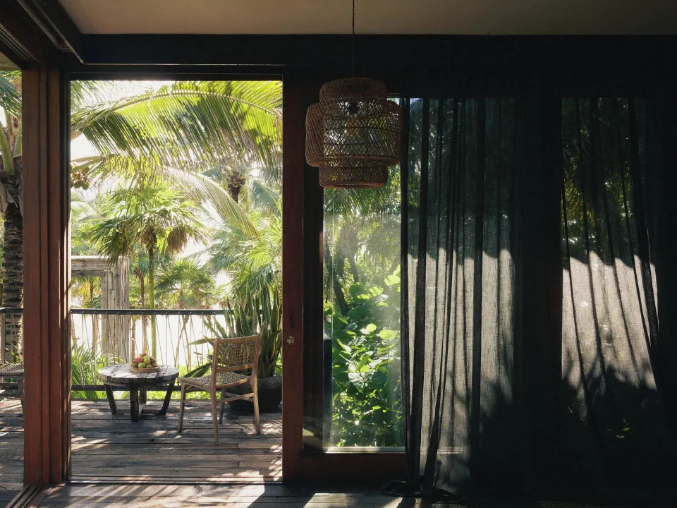 Terrace seating and palm foliage seen through sliding doors with sheer curtain at BE Destination Tulum