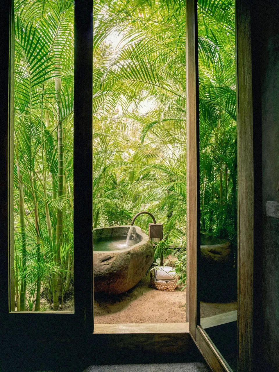 Outdoor stone bathtub surrounded by palms seen through a doorway at BE Destination Tulum.