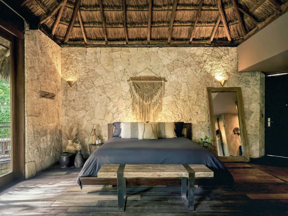 Palm Suite king bed with textured stone wall and thatched ceiling at BE Destination Tulum