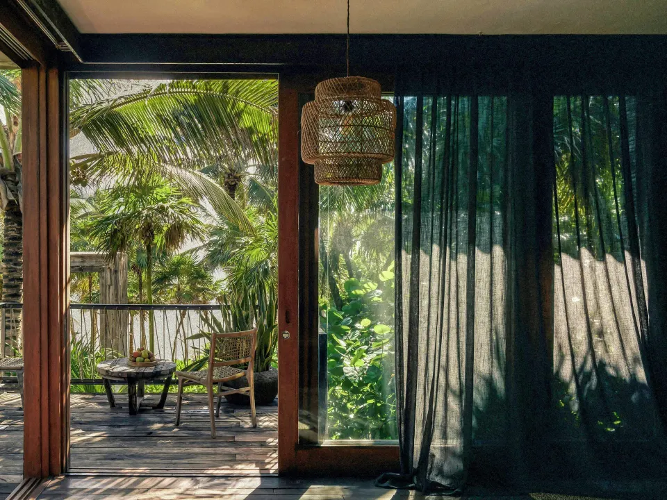 Sliding glass doors from suite opening to a shaded terrace with wicker chairs at BE Destination Tulum.