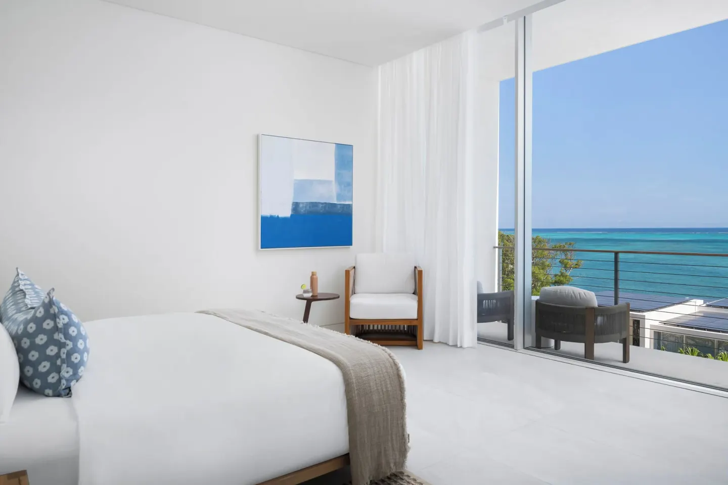 Ocean-view bedroom at Beach Enclave with a white bed, armchair, artwork, and balcony doors.