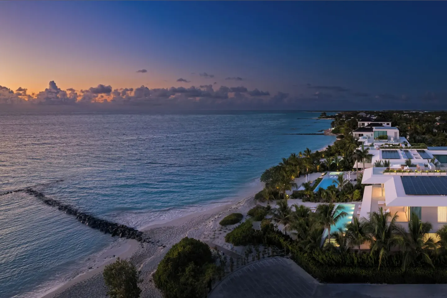 Sunset aerial of Beach Enclave villas along the coastline with pools, palms and the ocean at dusk.