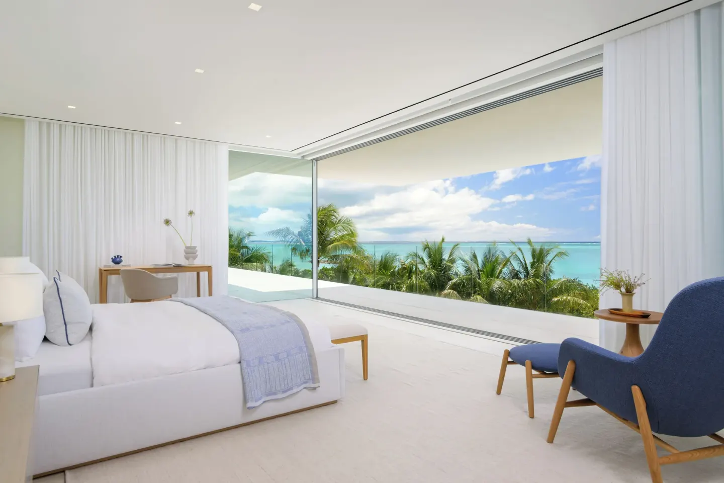 Oceanfront bedroom with a large bed, desk, blue chair and open glass wall at Beach Enclave.