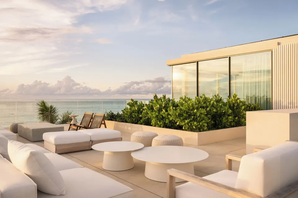 Sunlit terrace lounge at Beach Enclave with white seating, round tables, greenery and ocean beyond.