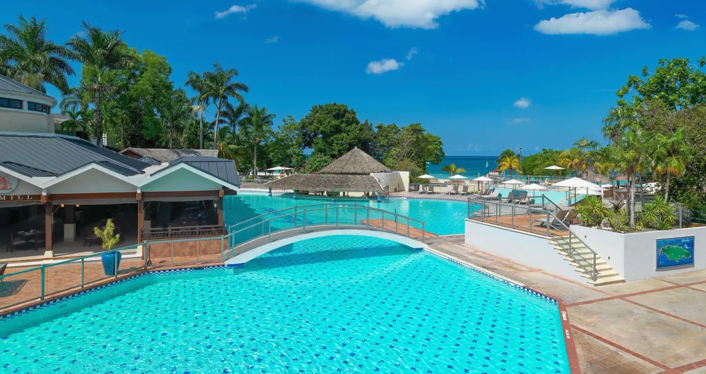 Large turquoise pool with bridge, loungers, and ocean beyond at Beaches Negril Resort.