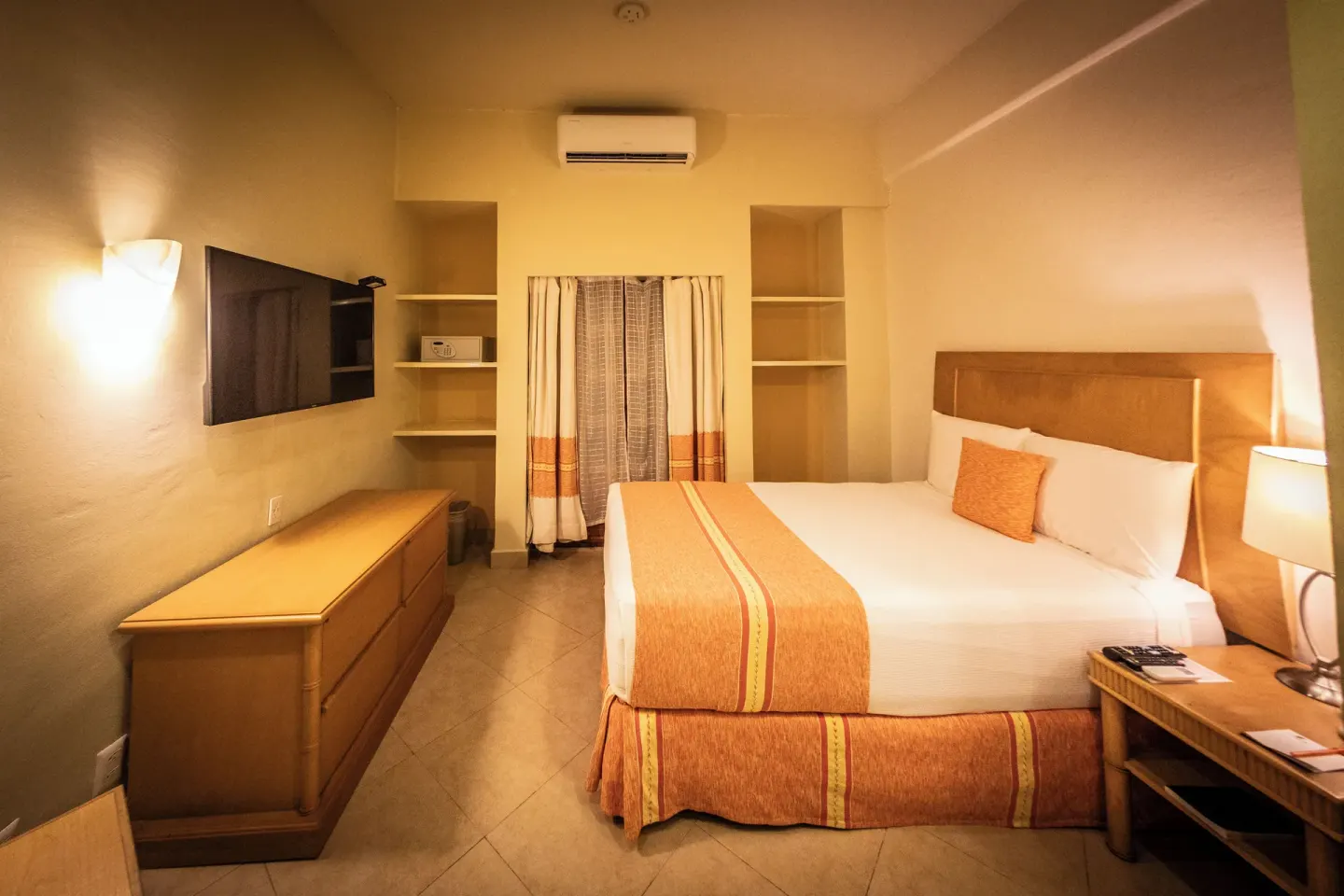 King bed guest room at Binniguenda Huatulco & Beach Club with wall TV, dresser, shelves, and window.