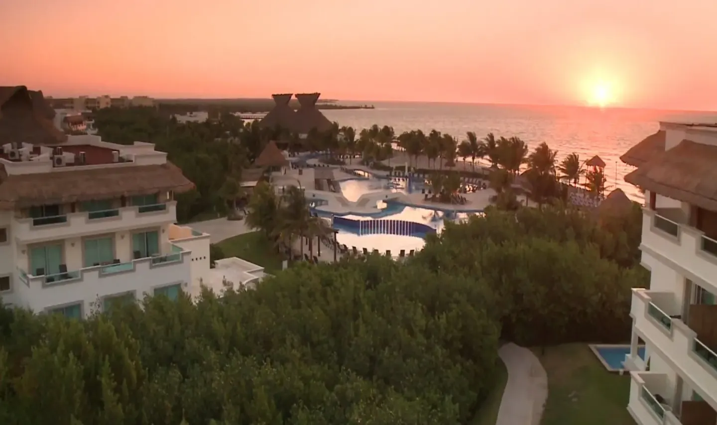 Sunset over pools, palms, guest buildings, and the sea at BlueBay Grand Esmeralda.