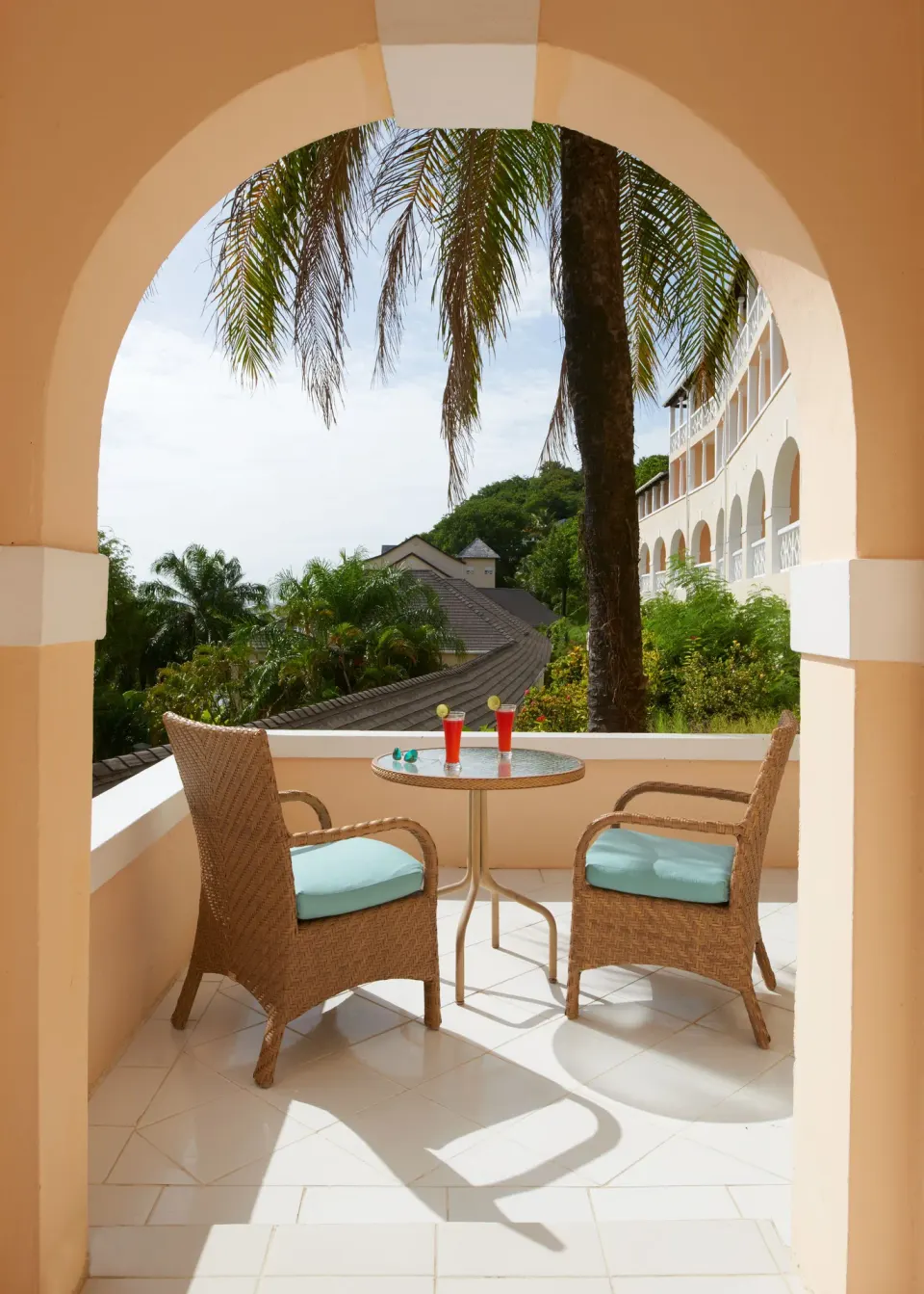 Arched BodyHoliday balcony with wicker chairs, a small table, and tropical garden views.