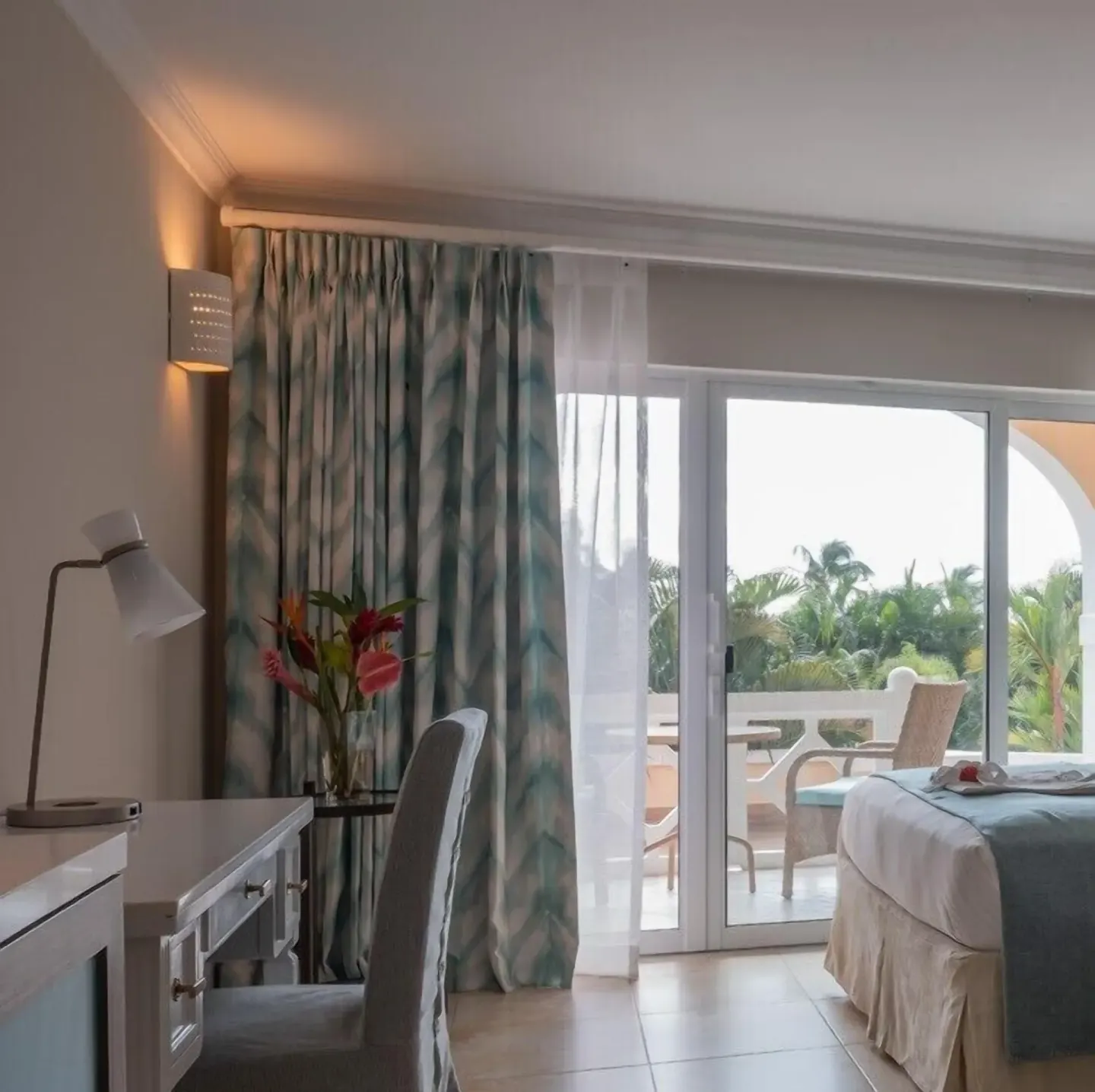 Guest room desk, balcony doors and bed at BodyHoliday with palms outside.