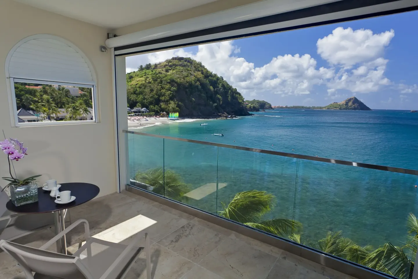 BodyHoliday balcony with a glass railing overlooking turquoise sea, beach, and green headlands.