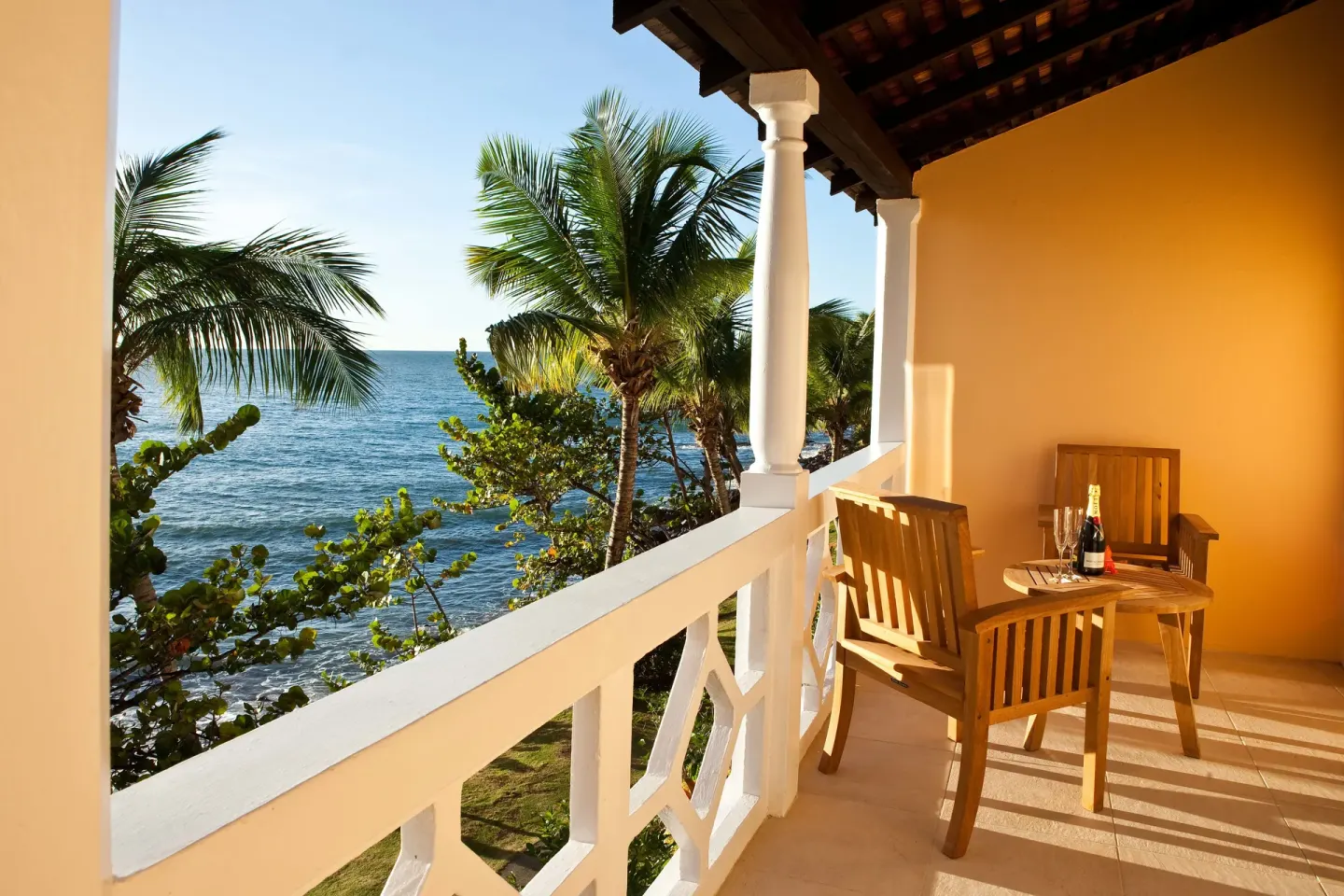 BodyHoliday oceanfront balcony with wooden chairs, a small table, palms, and sea views.