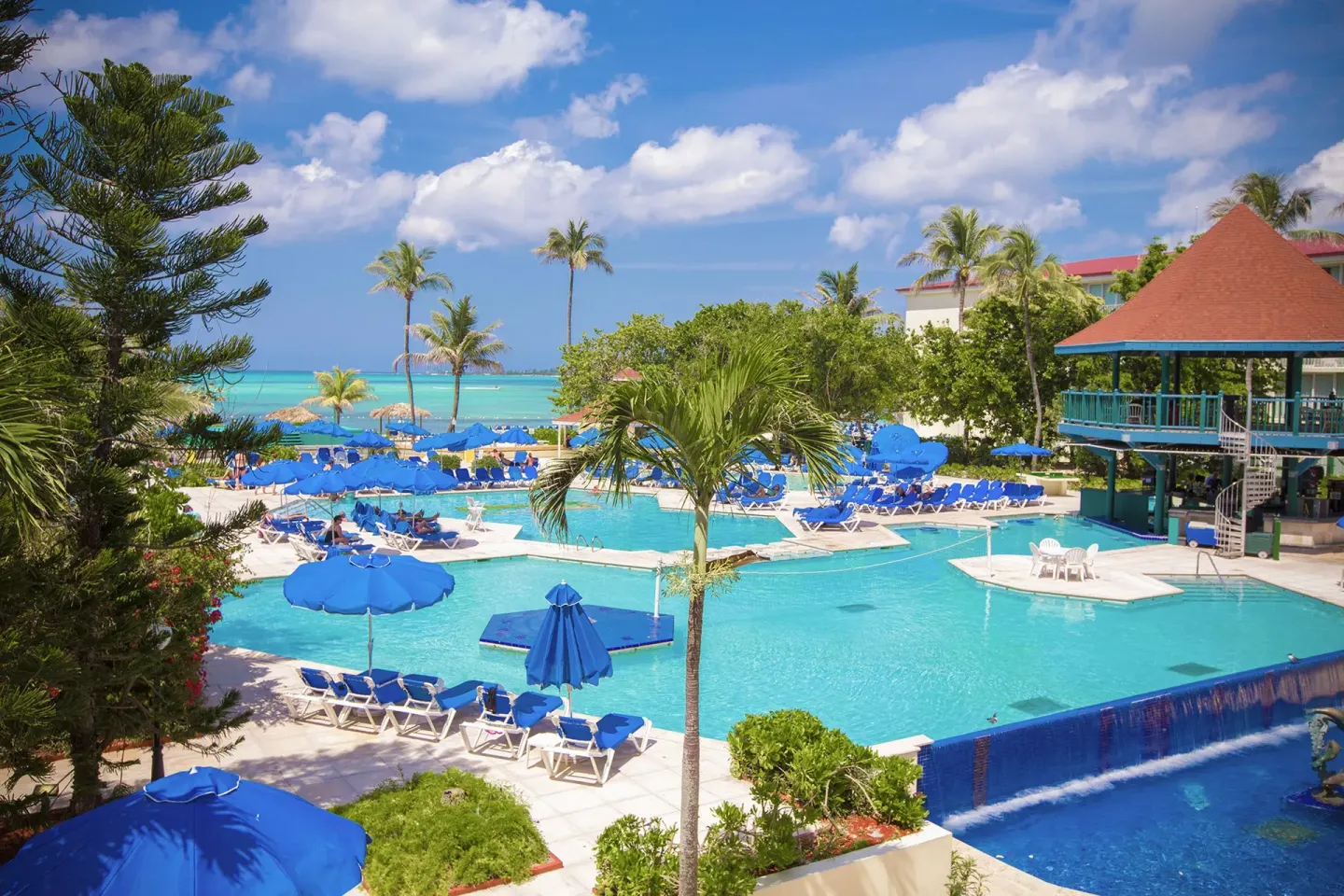Pool deck with blue umbrellas, loungers, palms, and ocean beyond at Breezes Resort Bahamas.