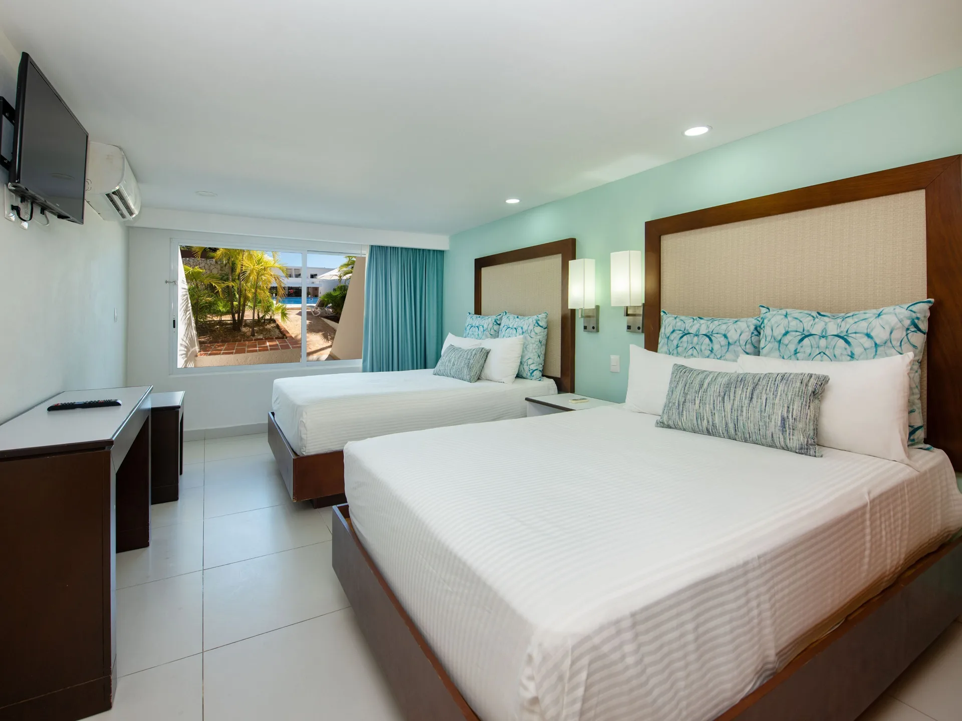 Standard Room at Cancun Bay Hotel with two beds, TV, desk and a window facing palms outside.