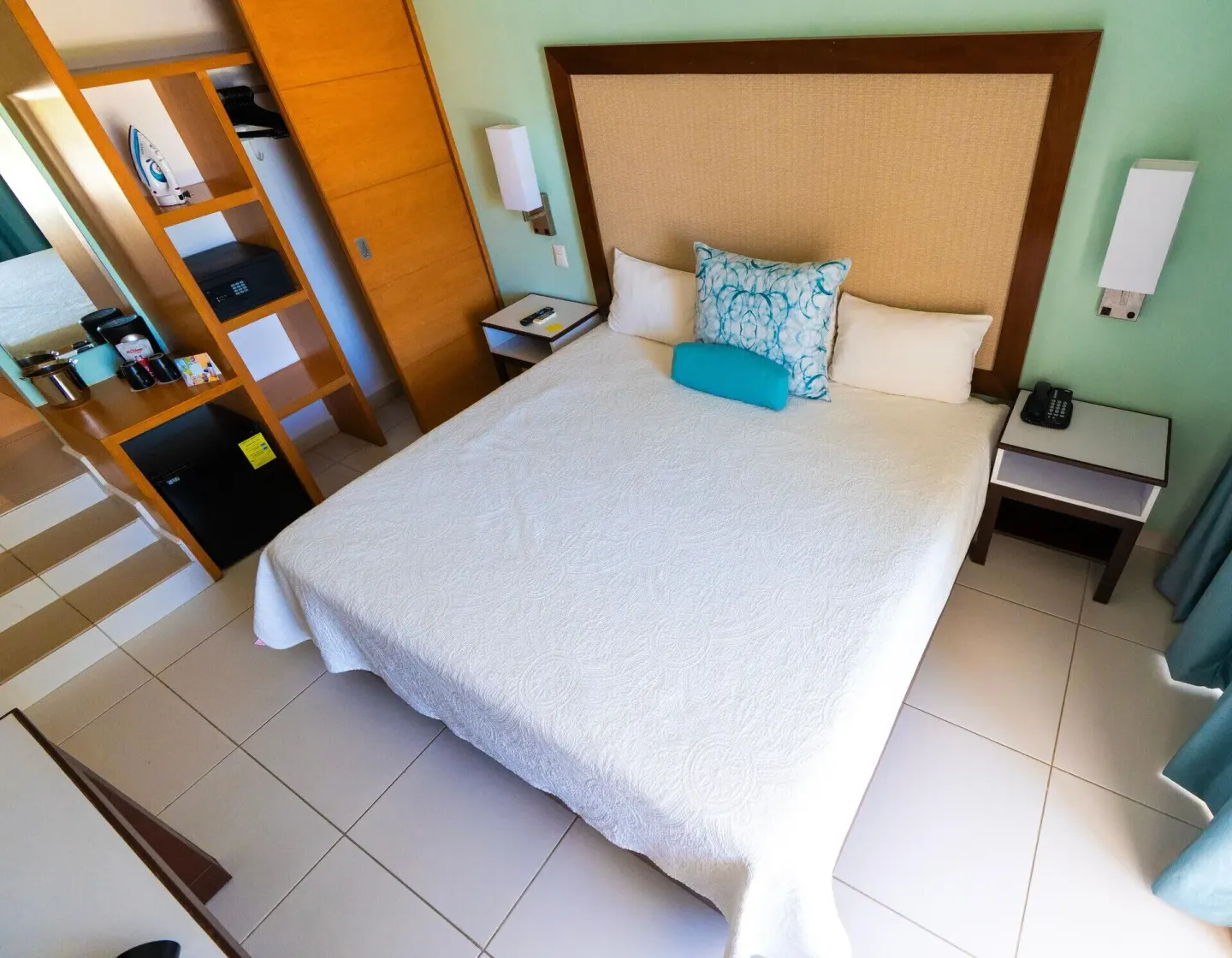 Standard Room king bed with wall lamps, open shelving, safe, and minibar at Cancun Bay.