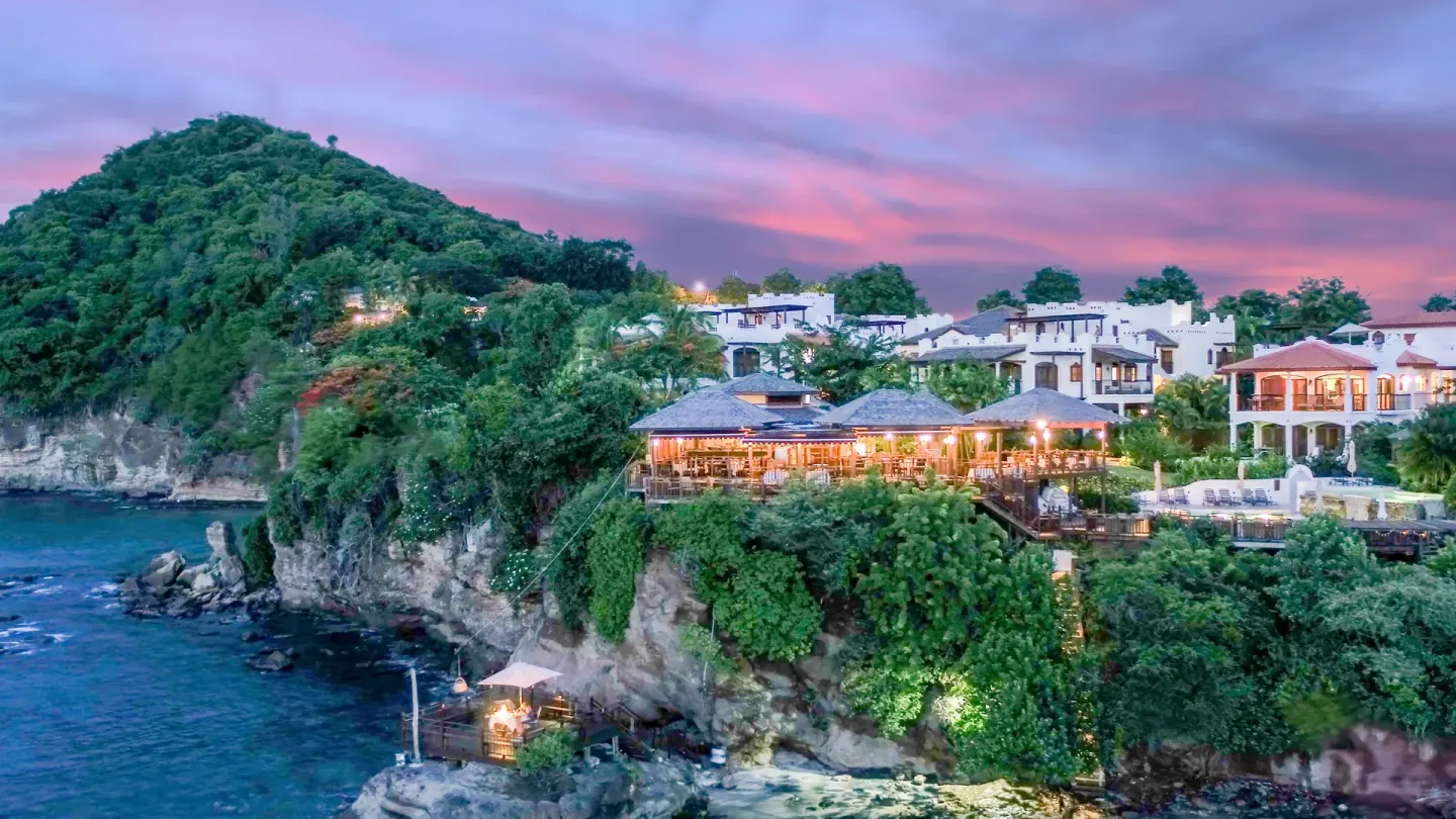 Sunset clifftop view of Cap Maison Resort & Spa with lights above the sea.