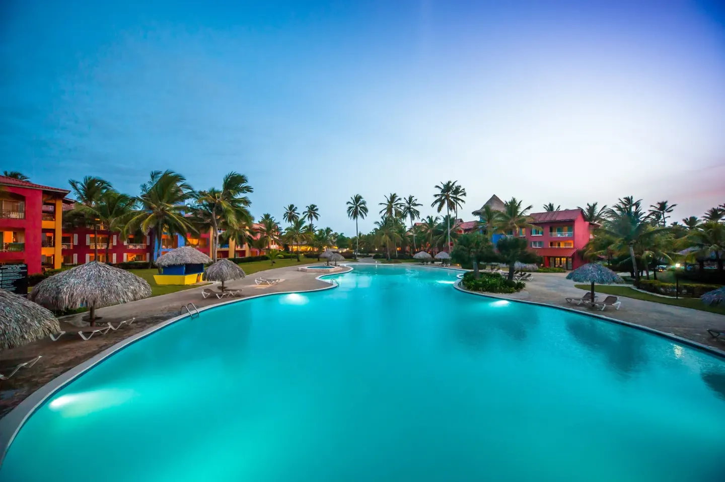 Evening view of a lit pool with palapas and colorful buildings at Caribe Deluxe Princess.