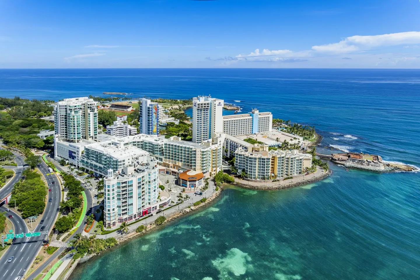 Aerial view of Caribe Hilton on a waterfront peninsula beside blue ocean.