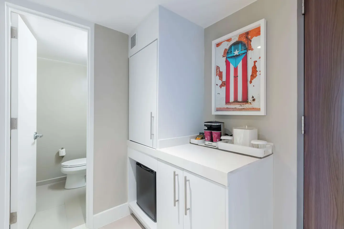Refreshment cabinet, mini-fridge, wall art, and restroom door in a Caribe Hilton guest room.