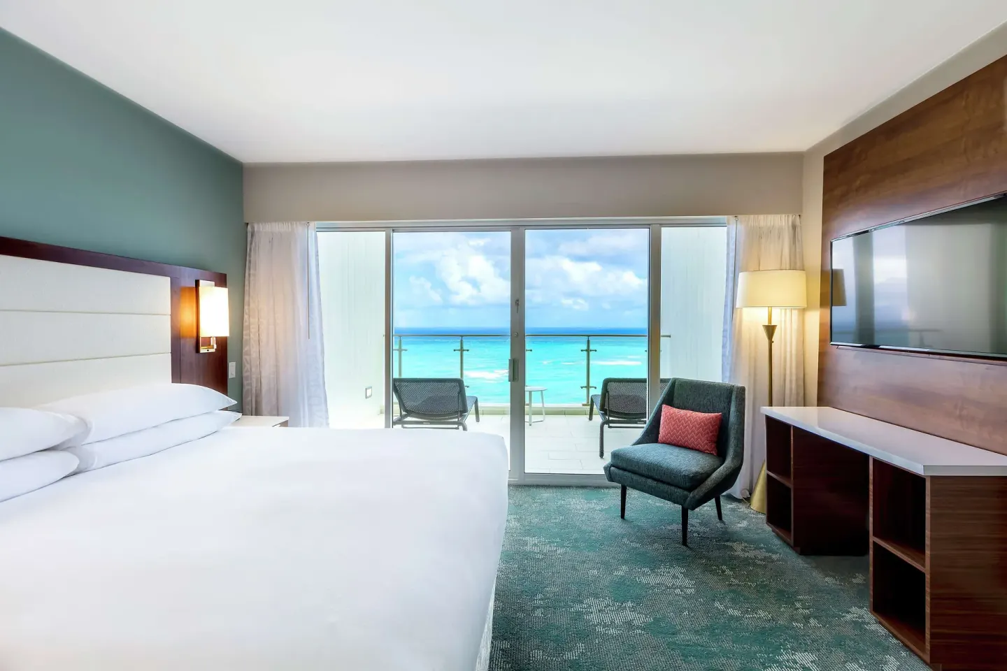 King bed, wall-mounted TV, chair, and ocean balcony in a suite at Caribe Hilton.