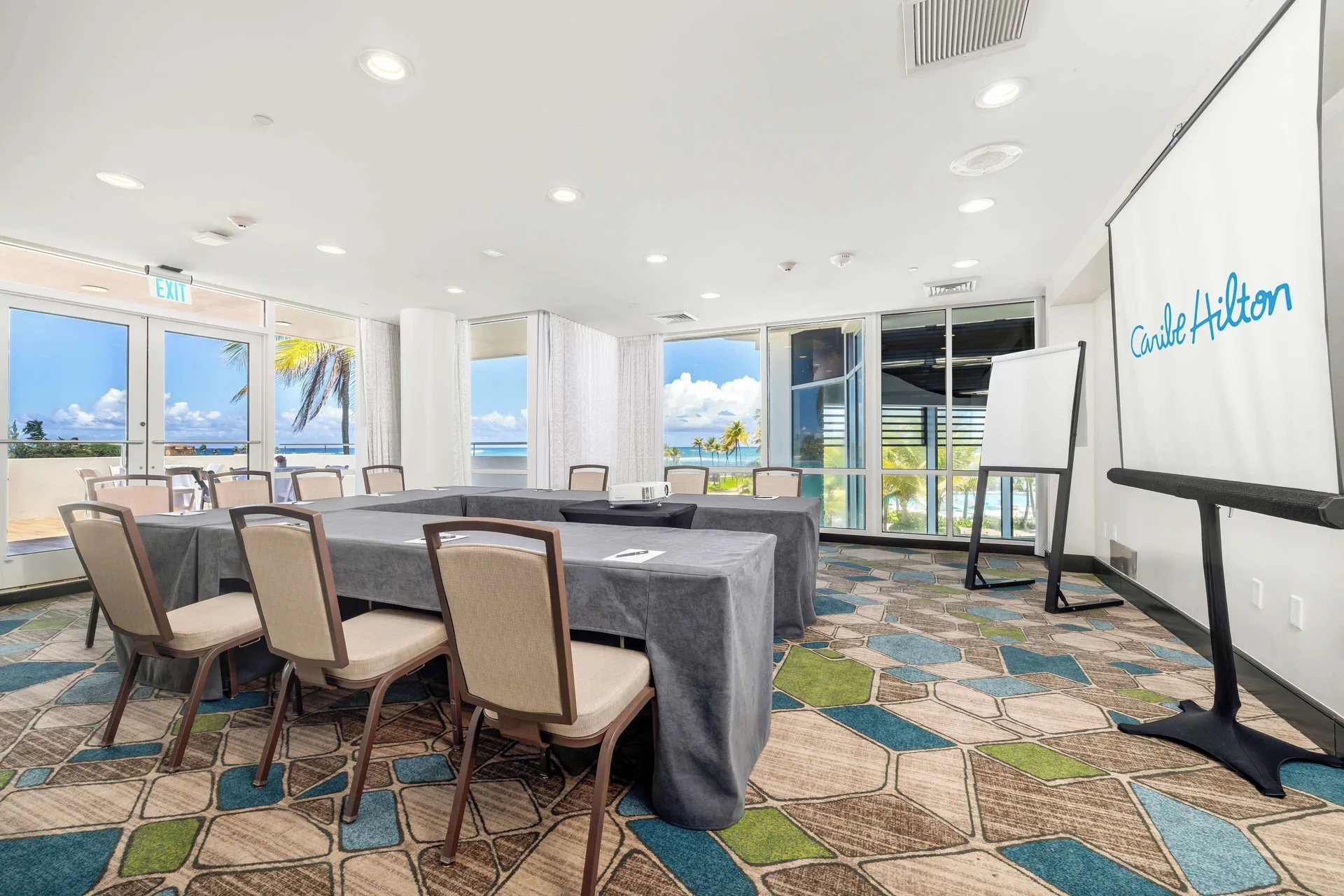 A meeting room with conference tables, ocean-facing windows, and a screen at Caribe Hilton.