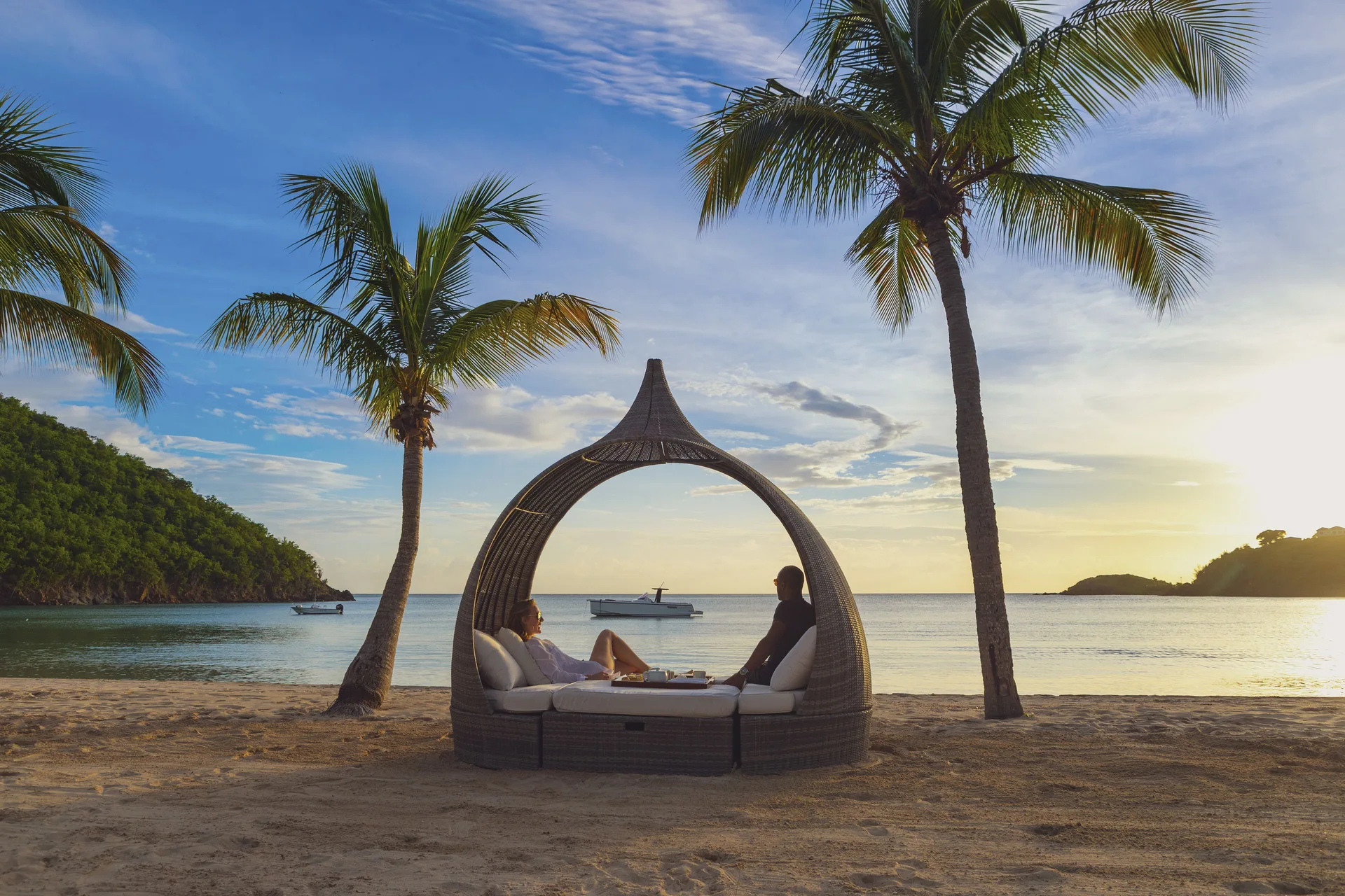 Guests relax in a woven beach cabana between palm trees at Carlisle Bay.