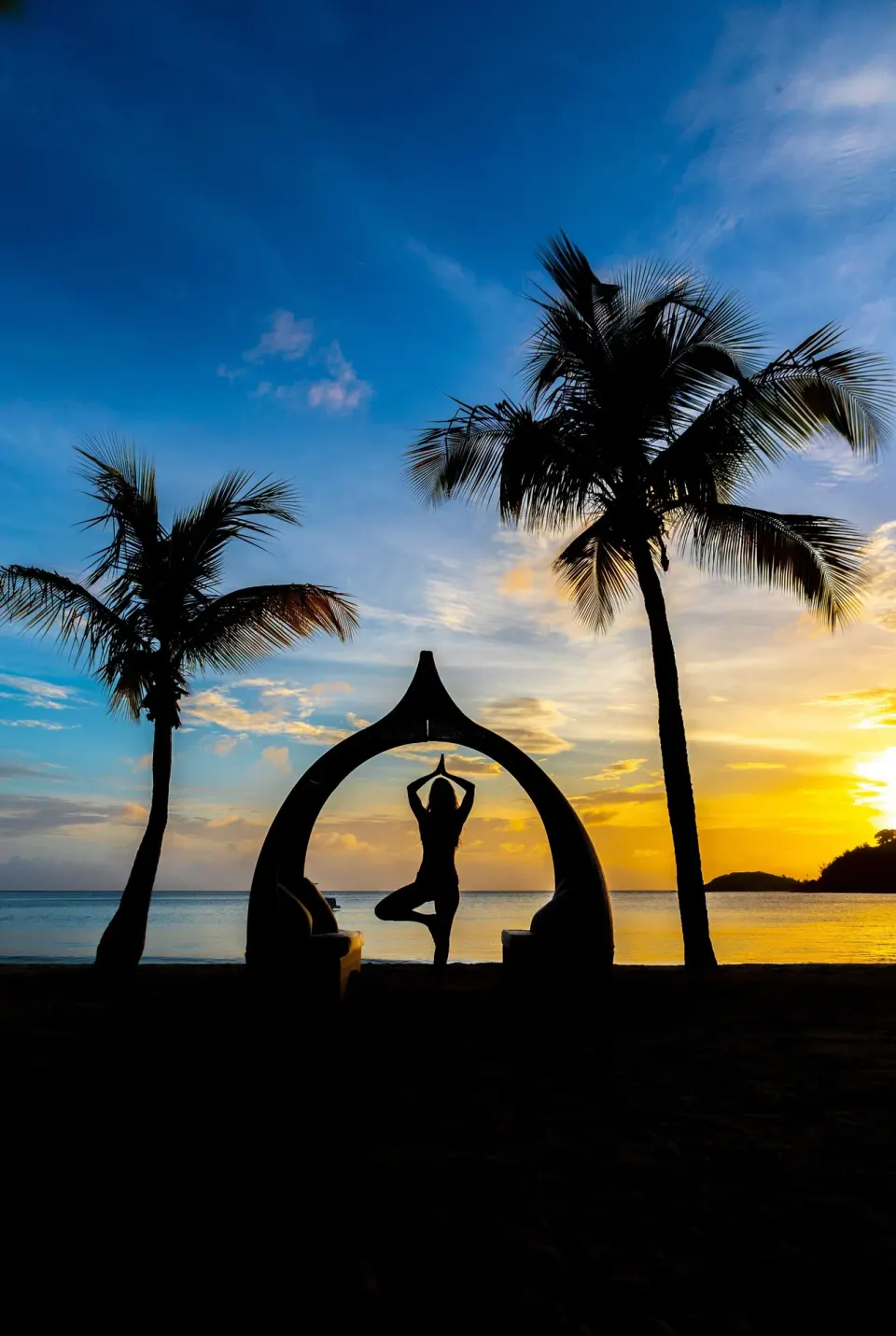 A yoga silhouette stands between palms on the beach at sunset at Carlisle Bay.