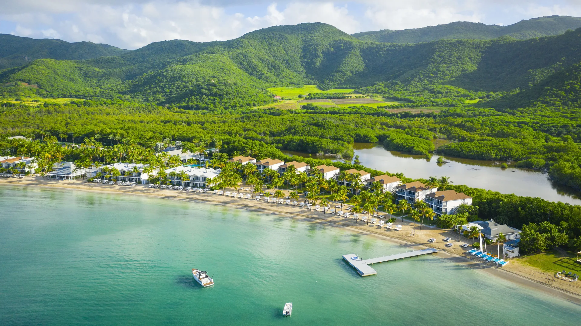 Wide aerial view of Carlisle Bay resort along the beach with green hills behind.