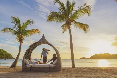 Guests relax in a beach daybed at sunset with a server at Carlisle Bay.