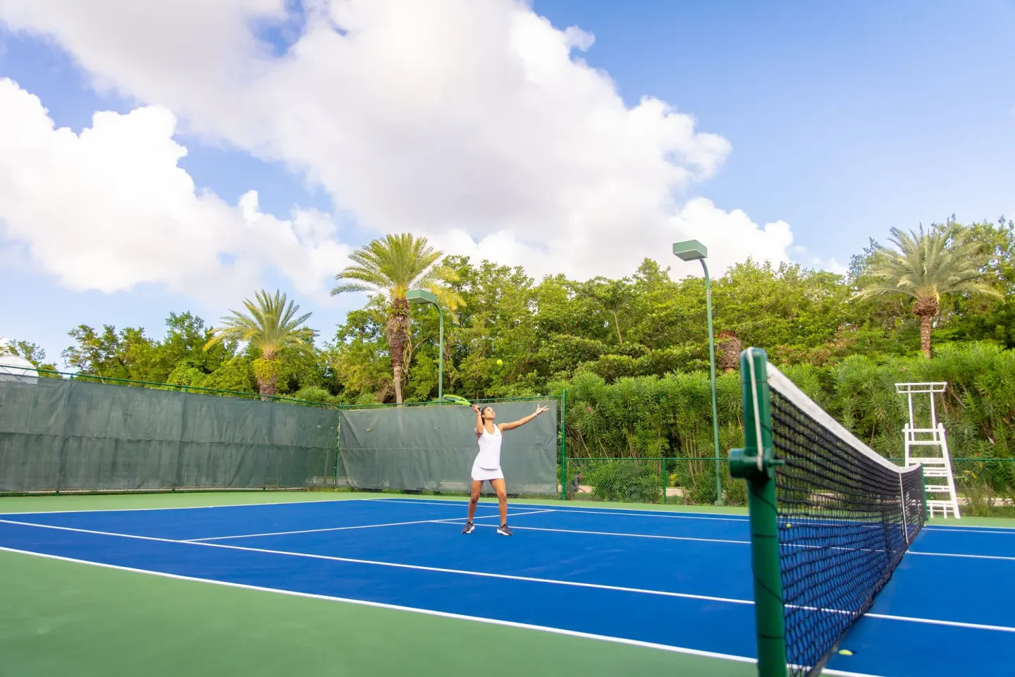 A player serves on a blue tennis court surrounded by greenery at Carlisle Bay.