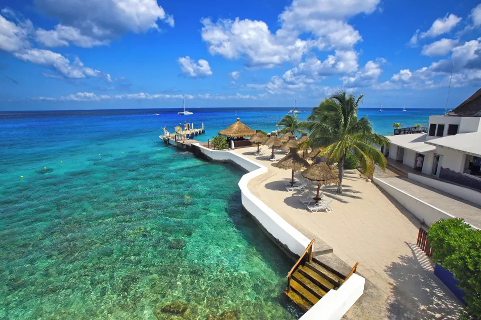 Resort beachfront with palapa umbrellas, dock and clear coral-blue water at Casa del Mar Cozumel Hotel & Dive Resort