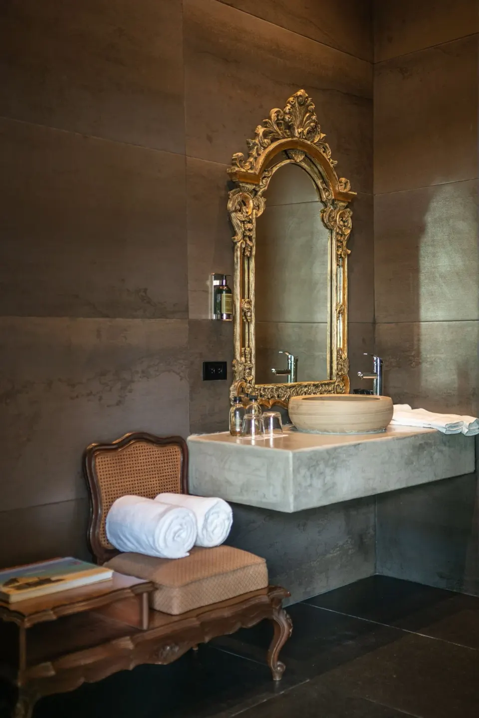 Ornate gold mirror above a stone sink and a bench with rolled towels in a Casa Malca suite.