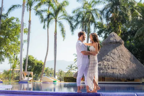 Couple standing by a palm-lined pool and thatched pavilion at Casa Velas.