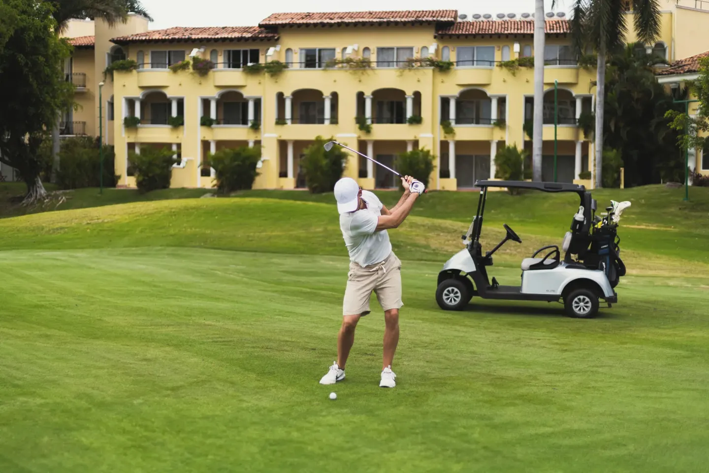 Golfer swinging on a green with a cart and resort building behind at Casa Velas.