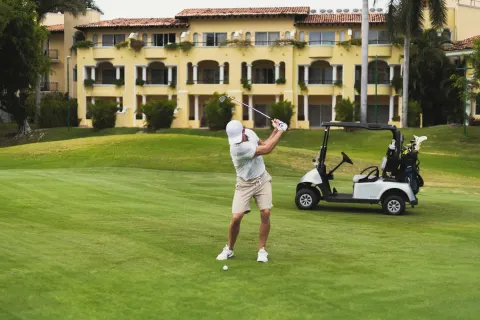 Golfer swinging on a green with a cart and resort building behind at Casa Velas.