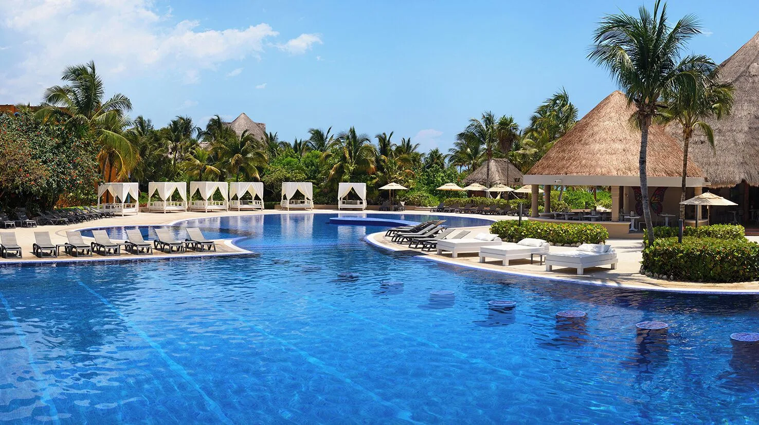 Large resort pool with loungers, cabanas, palms, and thatched roofs at Catalonia Playa Maroma.