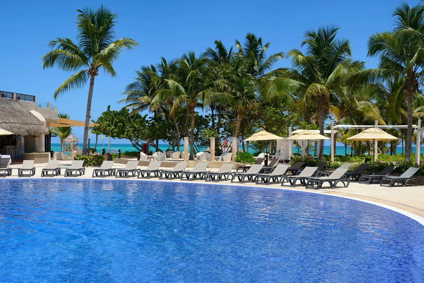 Curved pool with lounge chairs, palms, and the sea beyond at Catalonia Playa Maroma.